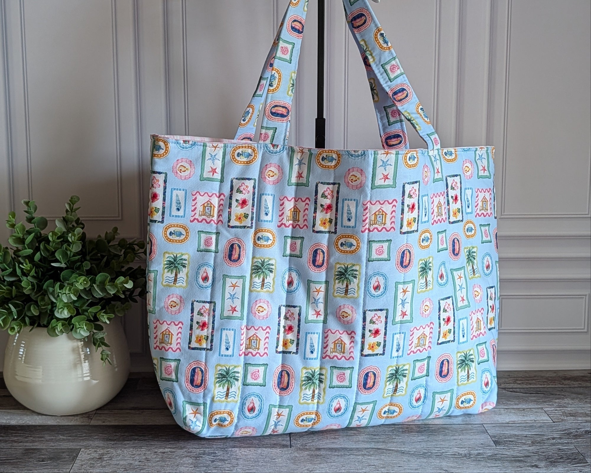 A light blue tote bag with a colorful patchwork print of palm trees, beach umbrellas, and resort-style icons, standing on a gray wooden floor next to a potted green plant.