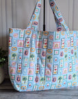 A light blue tote bag with a colorful patchwork print of palm trees, beach umbrellas, and resort-style icons, standing on a gray wooden floor next to a potted green plant.