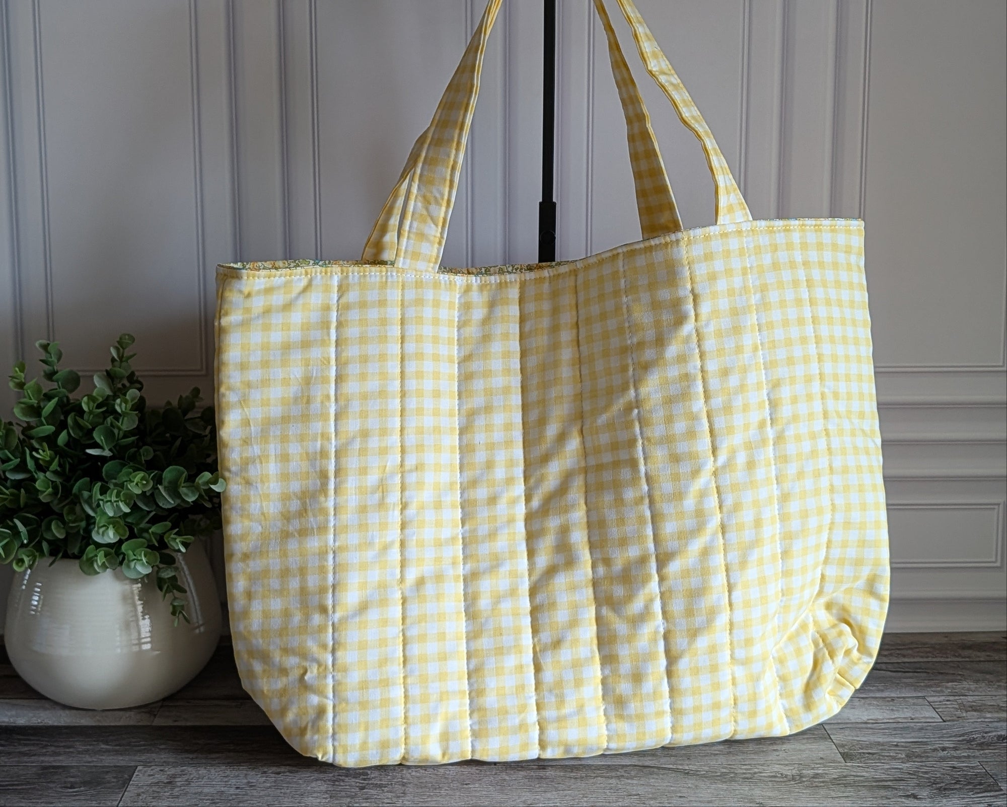 A quilted tote bag in a pale yellow and white gingham pattern, standing on a gray wooden floor next to a potted green plant.