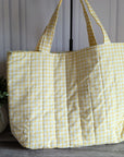 A quilted tote bag in a pale yellow and white gingham pattern, standing on a gray wooden floor next to a potted green plant.