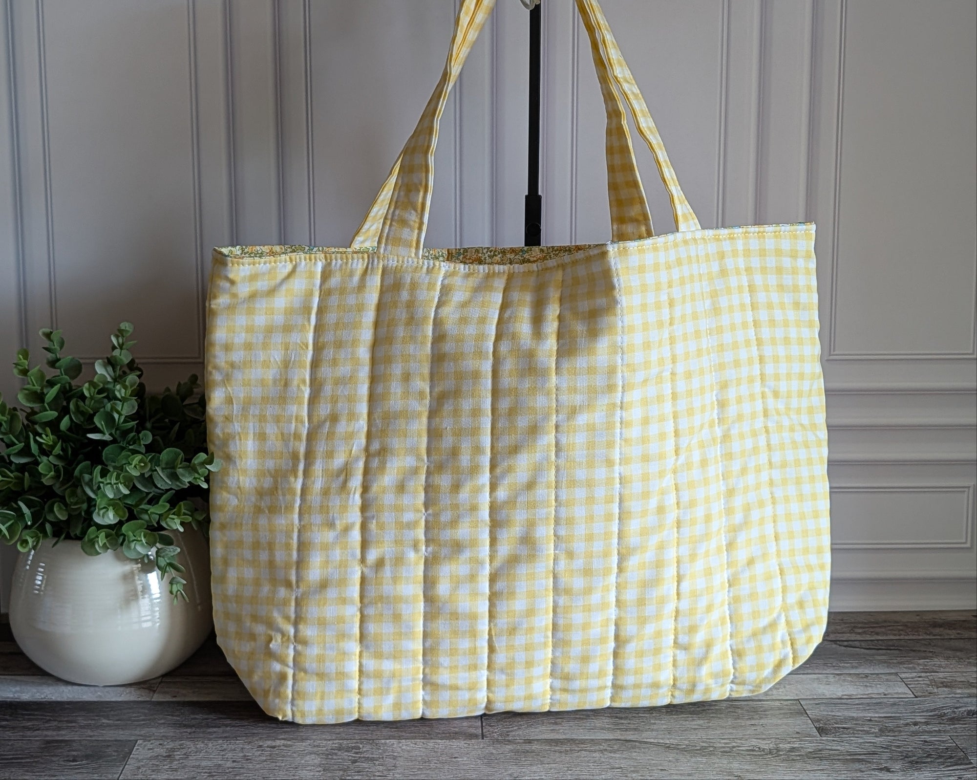 A large quilted tote bag in a pale yellow and white gingham check pattern, hanging on a black stand in front of a white paneled wall with a potted green plant on the left.