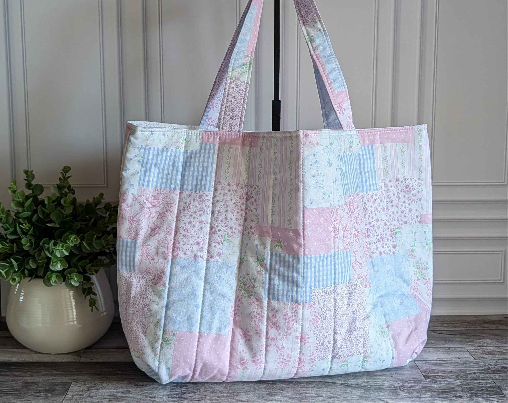A quilted patchwork tote bag in pastel pink, blue, and white fabrics, displayed on a stand in front of a white paneled wall with a potted plant on the left.