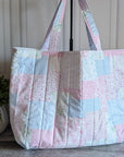 A quilted patchwork tote bag in pastel pink, blue, and white fabrics, displayed on a stand in front of a white paneled wall with a potted plant on the left.