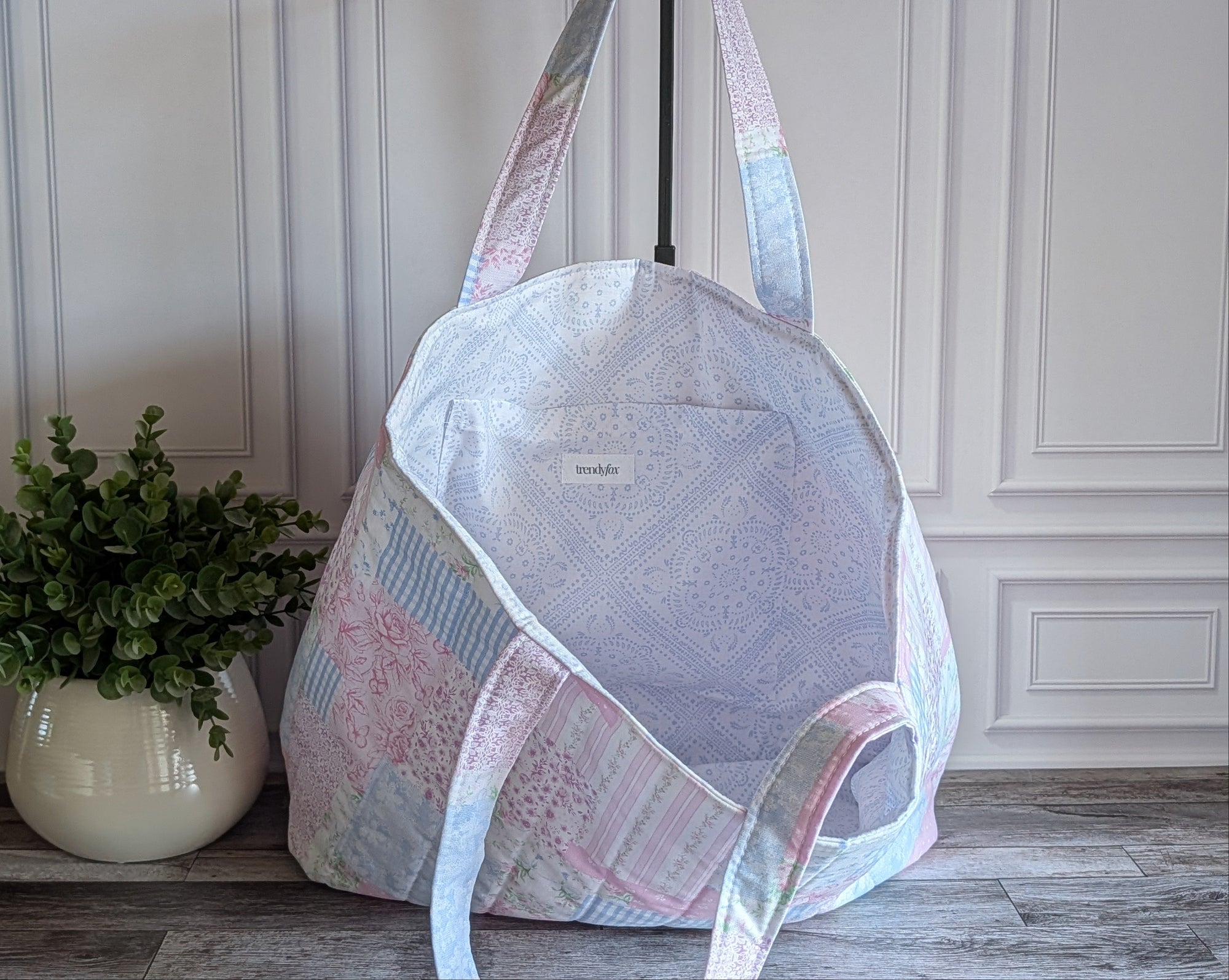A quilted patchwork tote bag in pastel pink, blue, and white fabrics, displayed on a stand in front of a white paneled wall with a potted plant on the left.