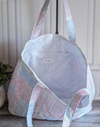 A quilted patchwork tote bag in pastel pink, blue, and white fabrics, displayed on a stand in front of a white paneled wall with a potted plant on the left.