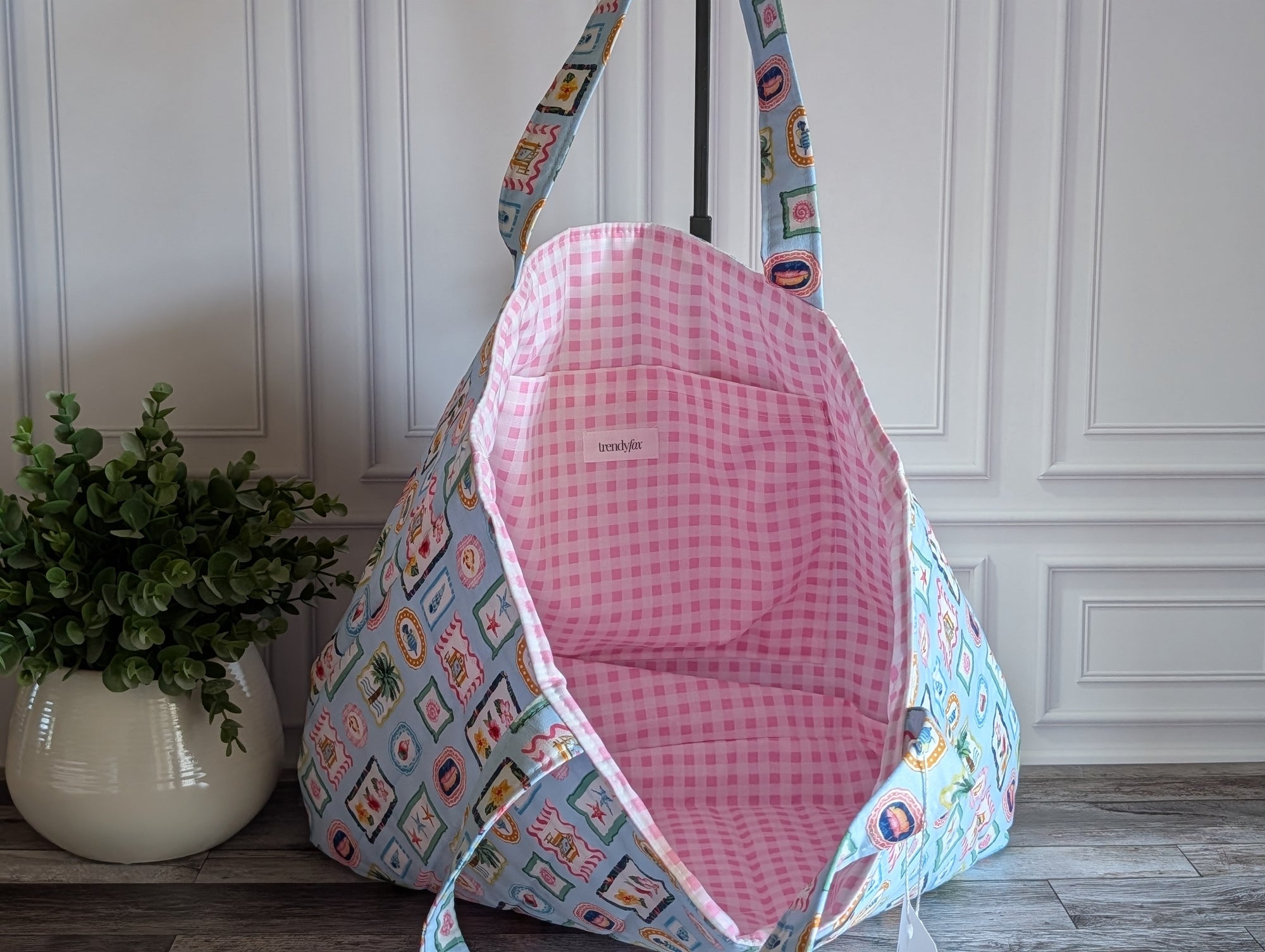 A fabric tote bag with a light blue and white patchwork print exterior and a pink gingham lining, shown open against a white paneled wall with a potted plant on the left.