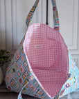 A fabric tote bag with a light blue and white patchwork print exterior and a pink gingham lining, shown open against a white paneled wall with a potted plant on the left.