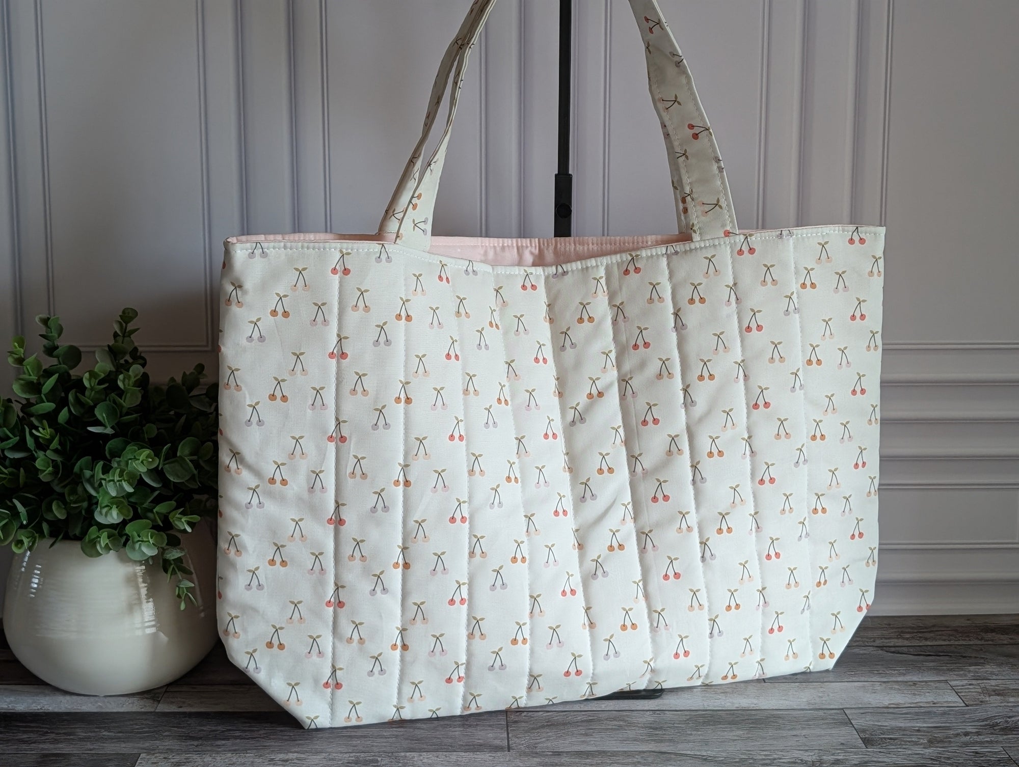 A quilted fabric tote bag with a repeating cherry print, standing on a dark wood floor against a paneled wall with a potted plant on the left.