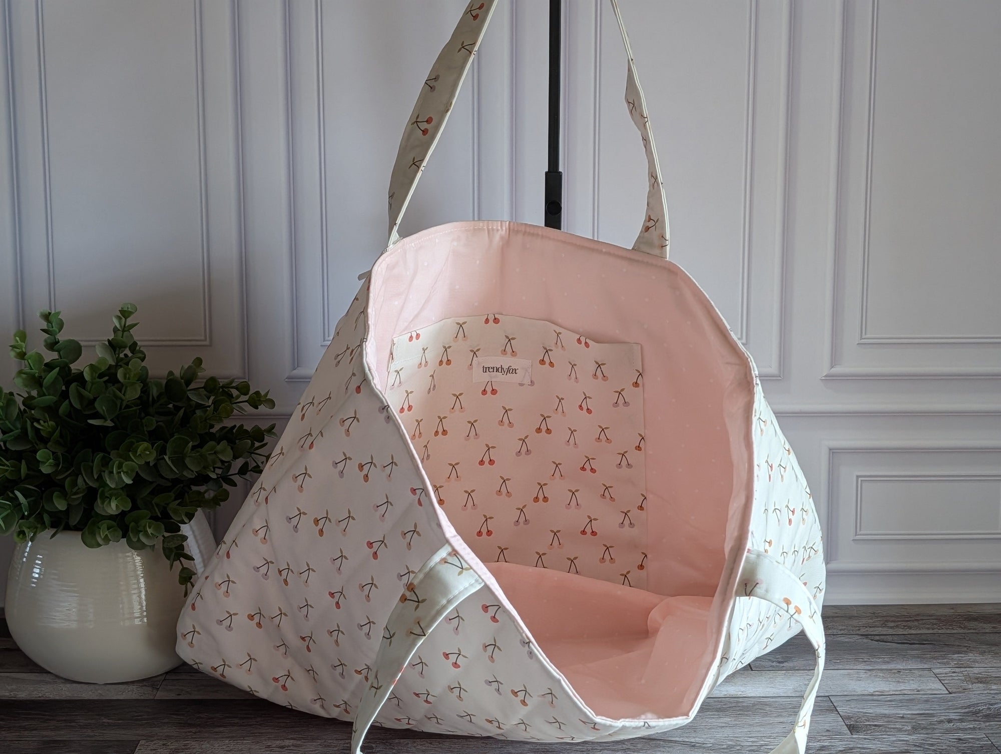 A quilted fabric tote bag with a repeating cherry print, standing on a dark wood floor against a paneled wall with a potted plant on the left.