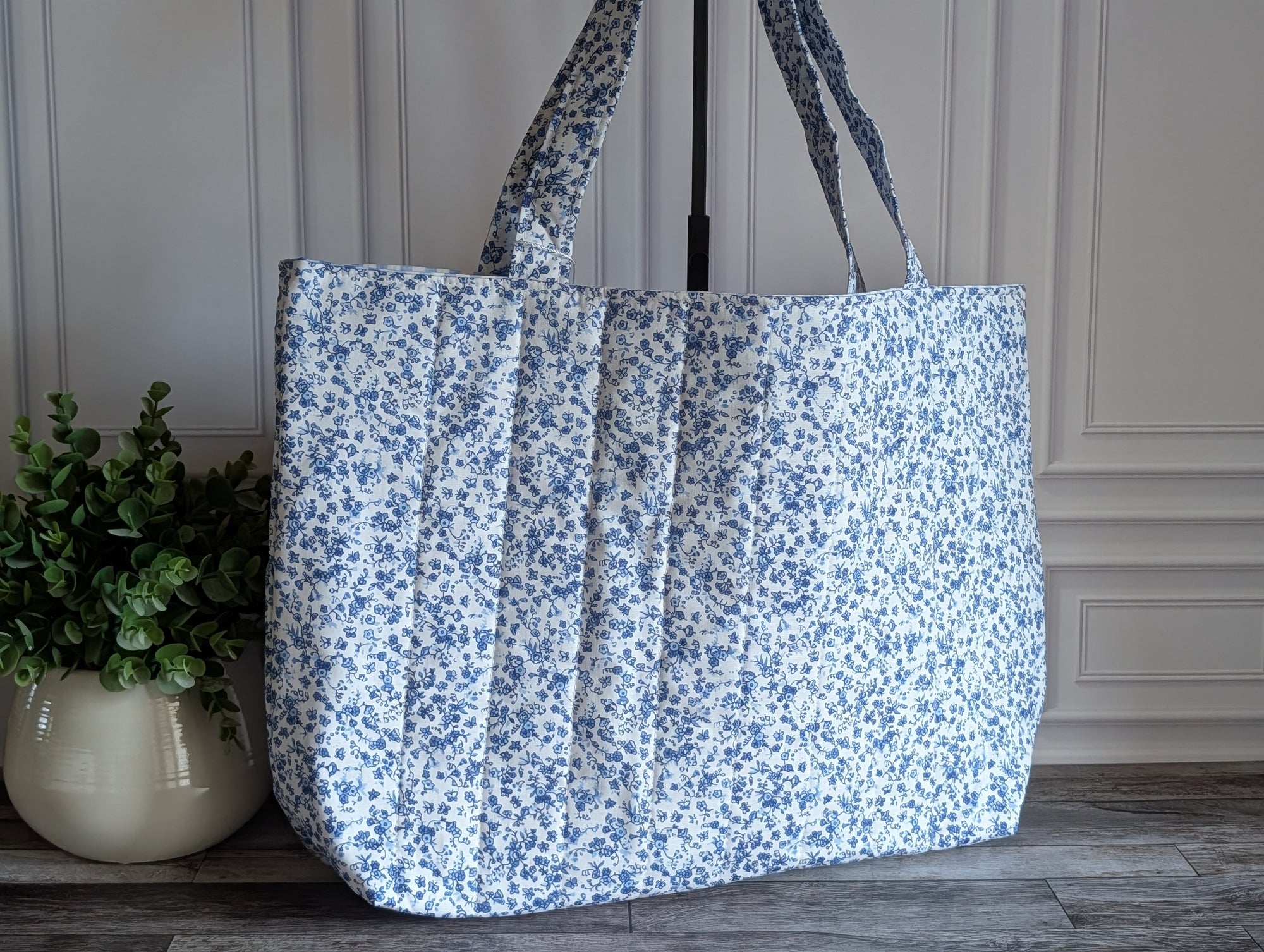 A large quilted tote bag with a blue and white floral print, standing on a wooden floor next to a potted green plant.