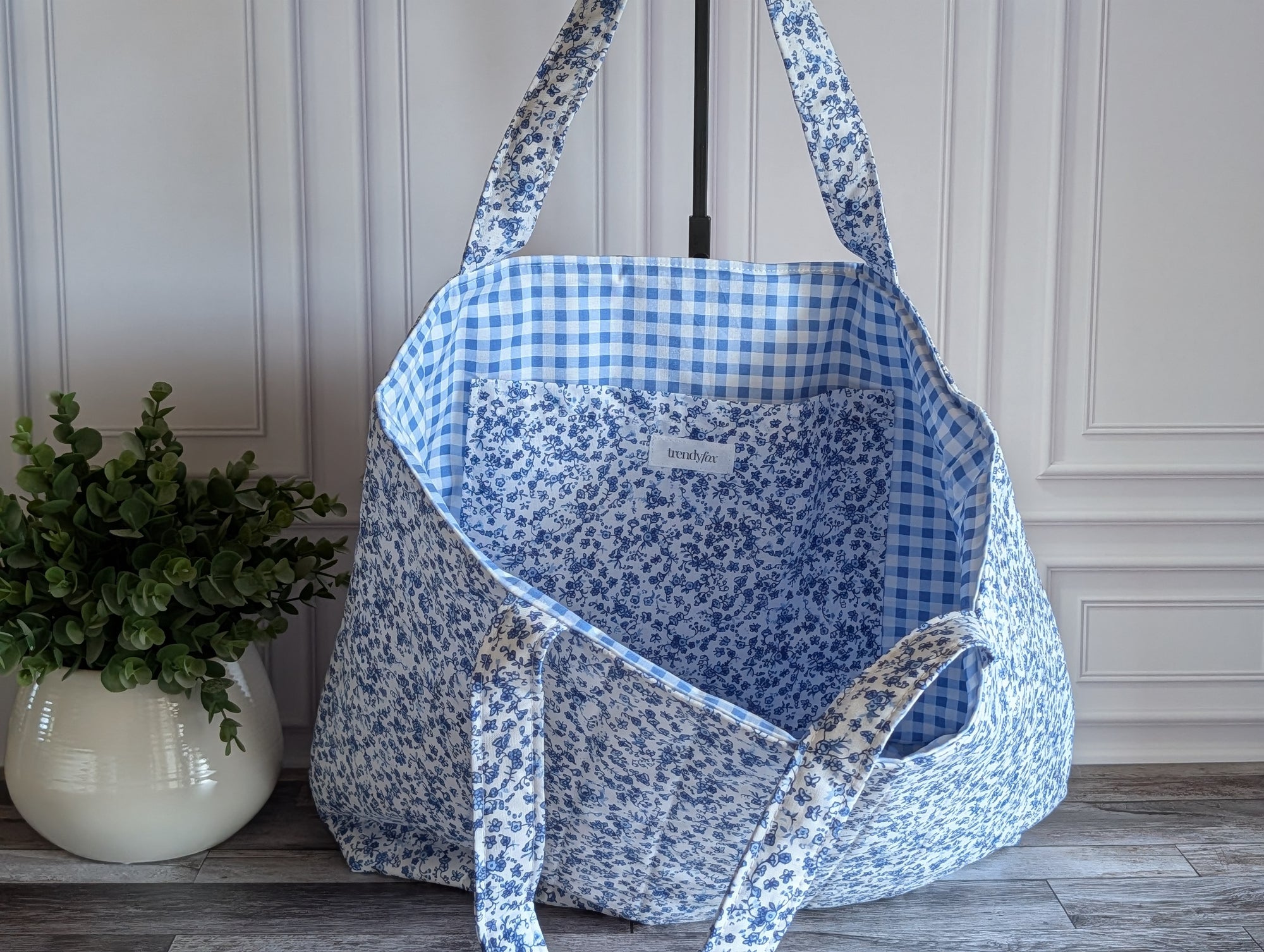 A large fabric tote bag with a blue and white floral print, standing on a gray wood floor next to a potted green plant against a paneled cream wall.