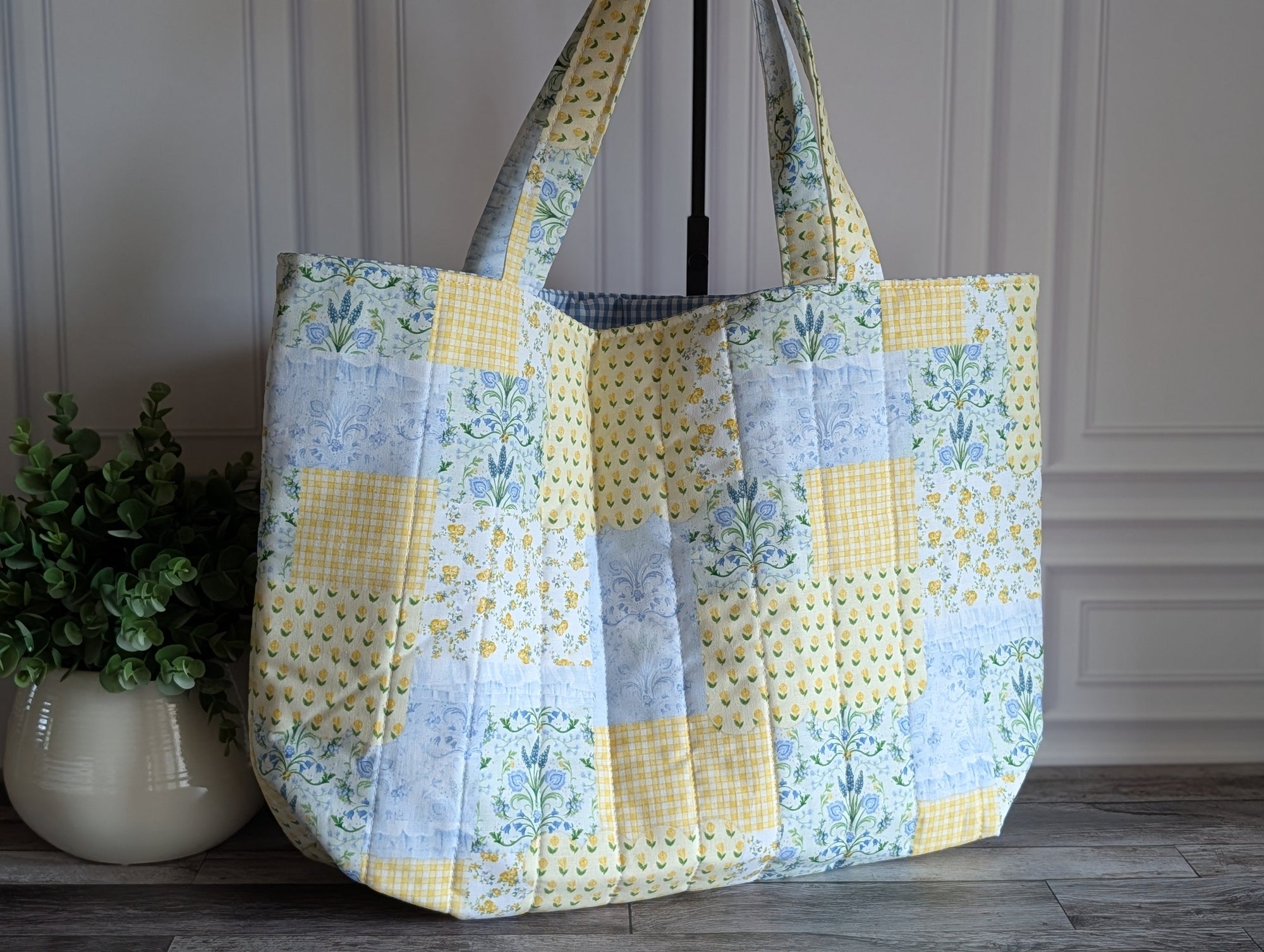 A large quilted patchwork tote bag with yellow, light blue, and white patterned fabric panels, featuring two shoulder straps, displayed on a wooden floor next to a potted plant.