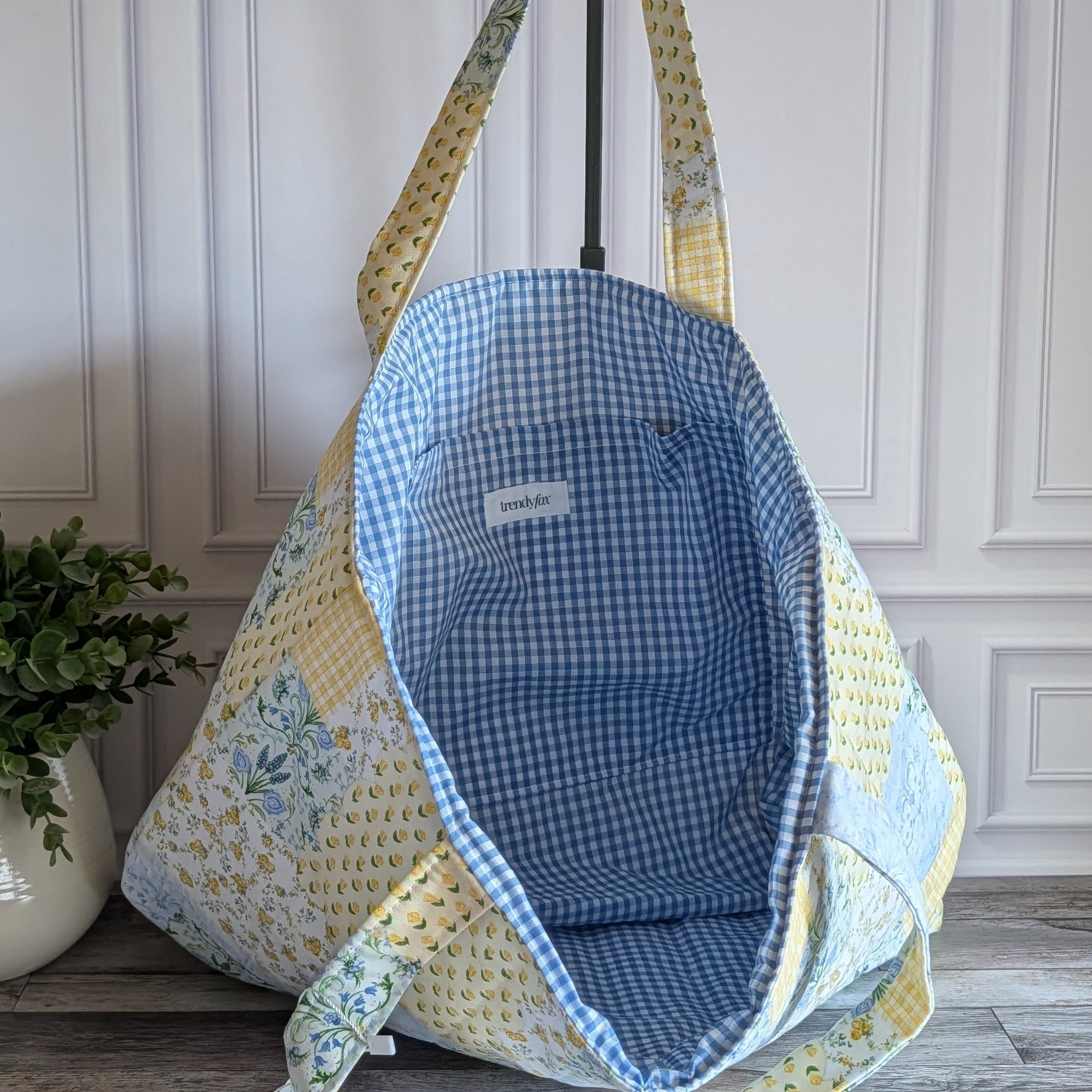 A quilted tote bag with a blue and white gingham interior and a yellow, white, and blue floral patchwork exterior, hanging on a black stand in front of a white paneled wall.