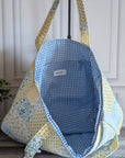 A quilted tote bag with a blue and white gingham interior and a yellow, white, and blue floral patchwork exterior, hanging on a black stand in front of a white paneled wall.