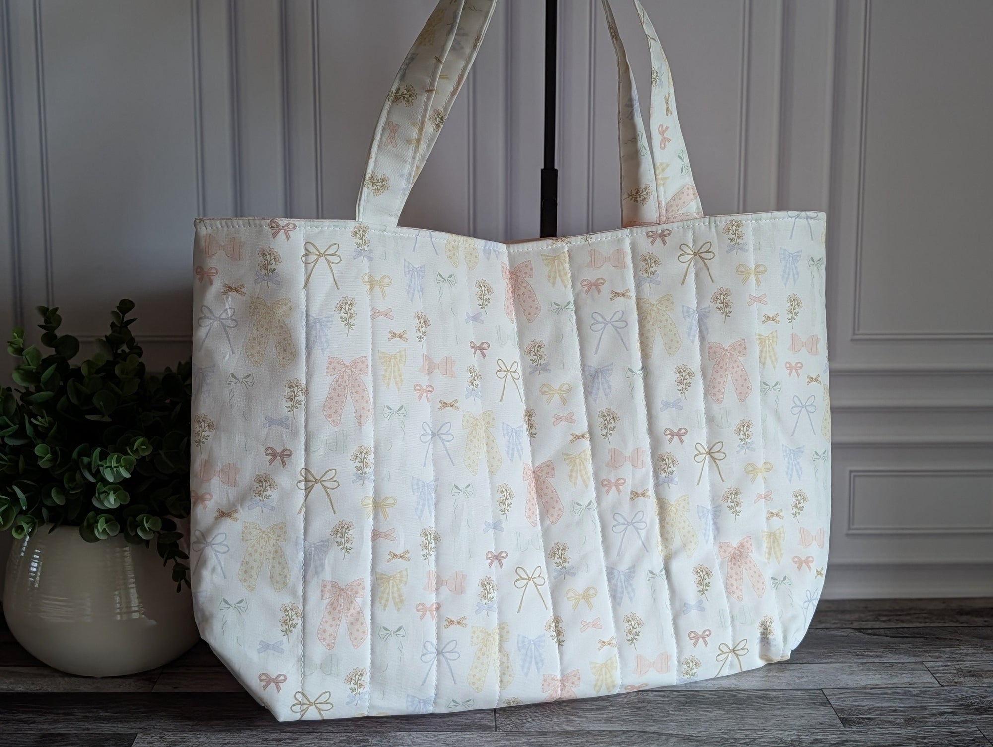A large quilted fabric tote bag with a delicate pastel floral and bow pattern, standing on a dark wood floor next to a potted green plant.