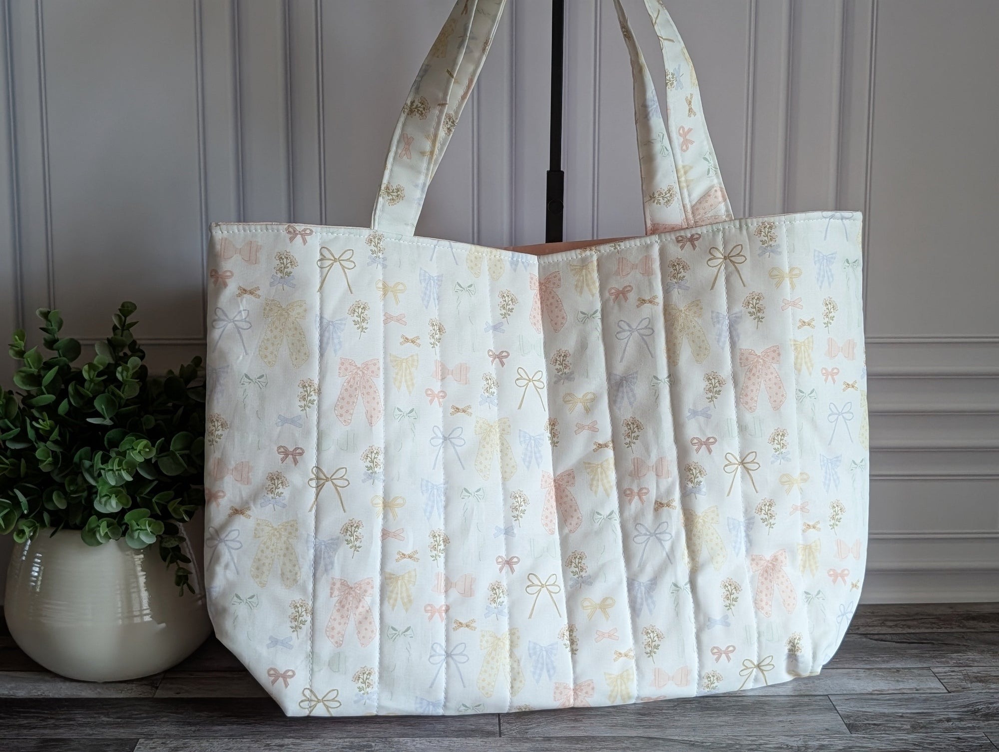 A large quilted fabric tote bag with a delicate pastel floral and bow pattern, standing on a dark wood floor next to a potted green plant.