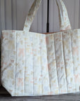 A large quilted fabric tote bag with a delicate pastel floral and bow pattern, standing on a dark wood floor next to a potted green plant.
