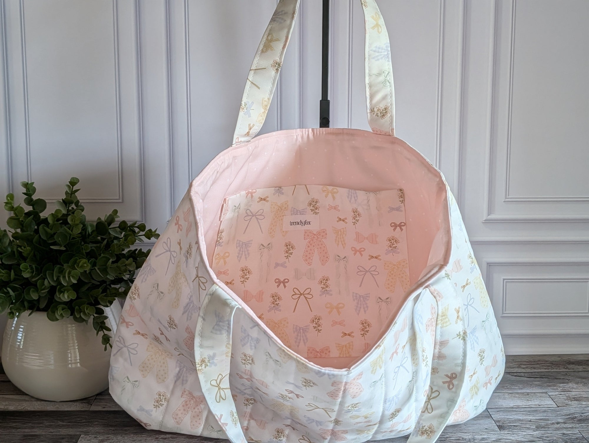 A large quilted fabric tote bag with a delicate pastel floral and bow pattern, standing on a dark wood floor next to a potted green plant.