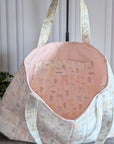 A large quilted fabric tote bag with a delicate pastel floral and bow pattern, standing on a dark wood floor next to a potted green plant.